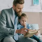 businessman and little son reading book together an sofa at home, work and life balance